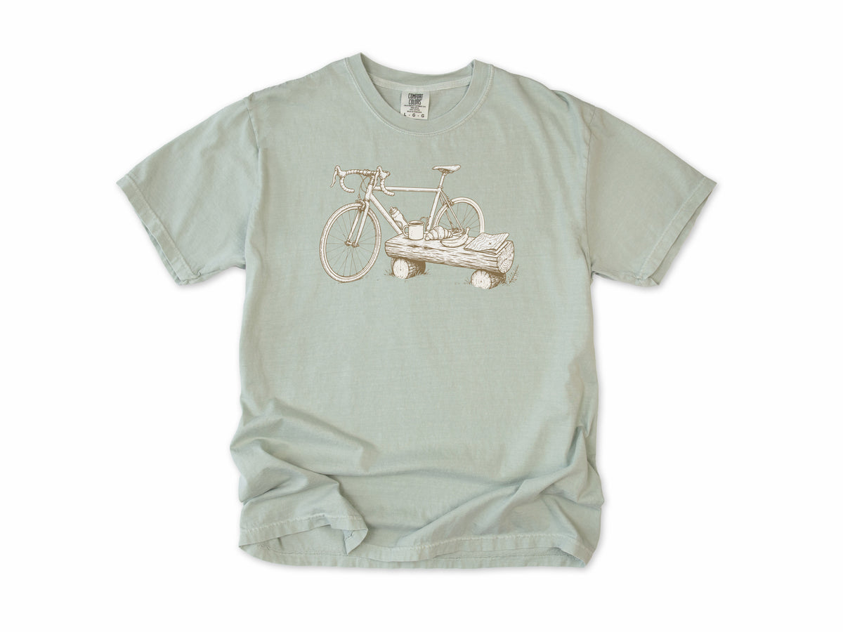 Funny cycling T-shirt in Bay, a bike propped on a log with a drink, for gravel and trail riders