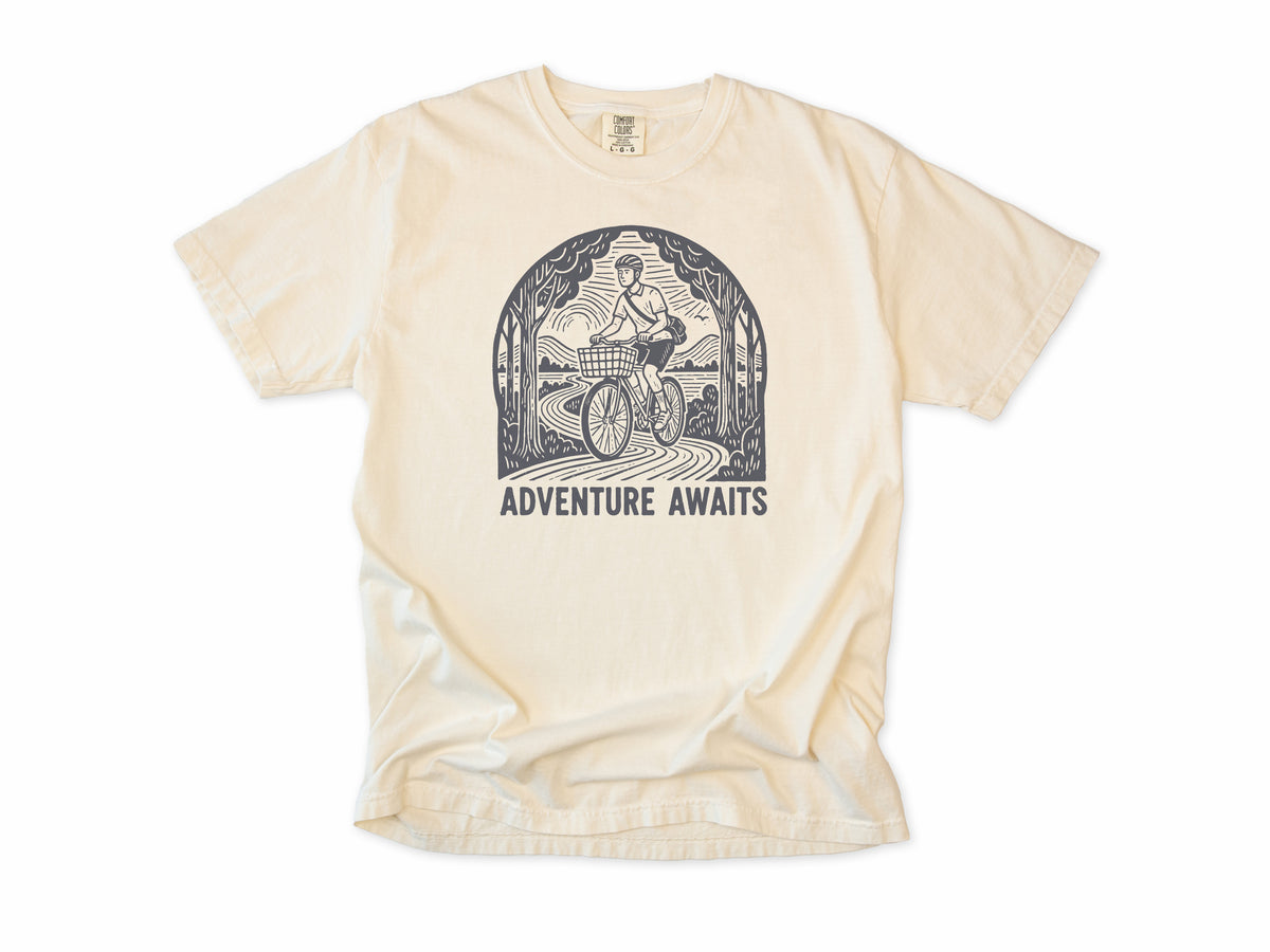 Cycling T-shirt in Ivory, a woodcut cyclist on a country road, 'Adventure Awaits', for touring and leisure cyclists