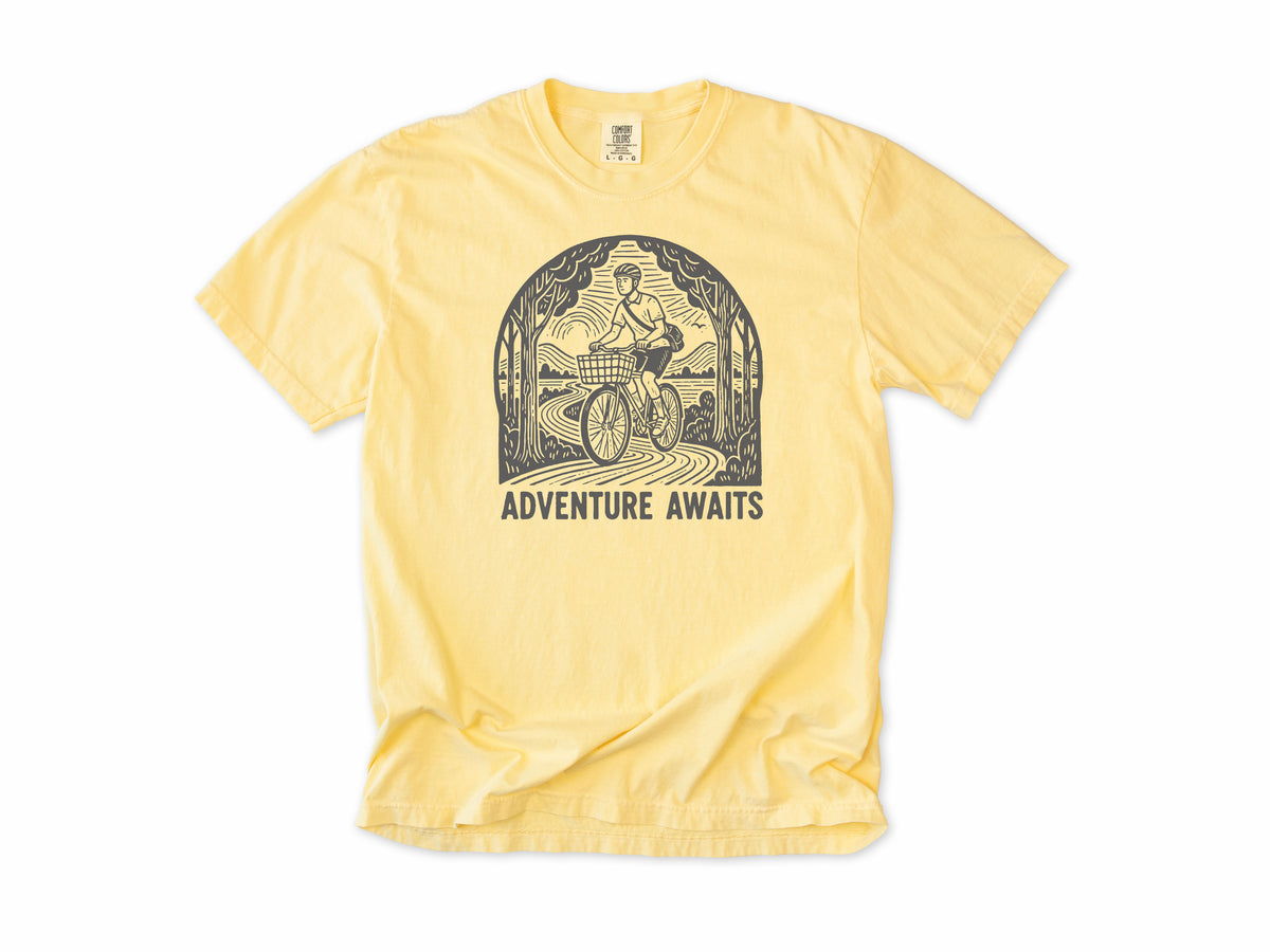 Cycling T-shirt in Butter, a woodcut cyclist on a country road, 'Adventure Awaits', for touring and leisure cyclists